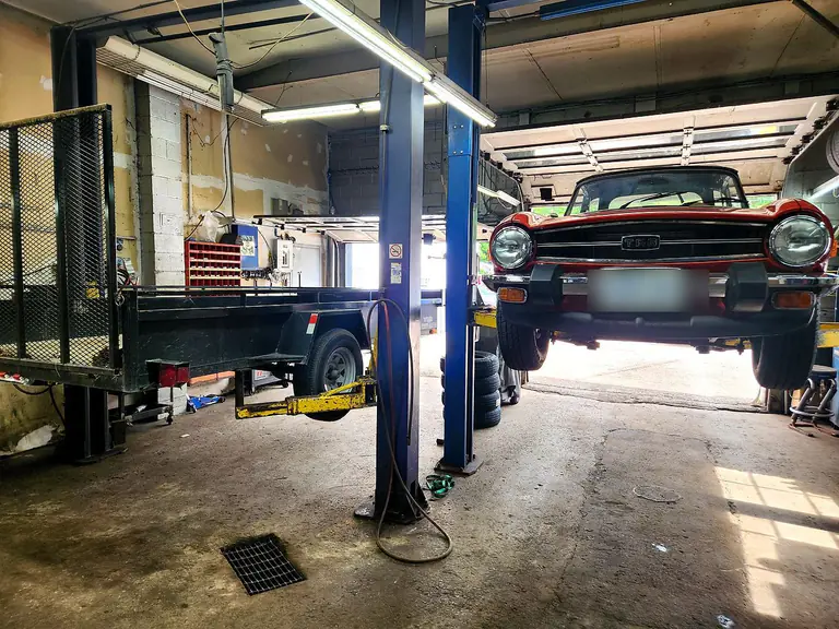 A classic car up on the hoist beside a flatbed trailer, looking down the length of the workshop